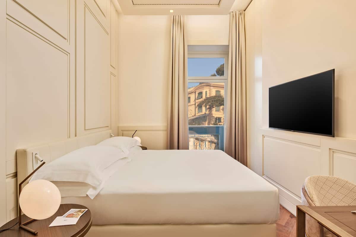 A hotel room with a bed, a TV, a bedside table with a lamp, and a view of a building through a window.