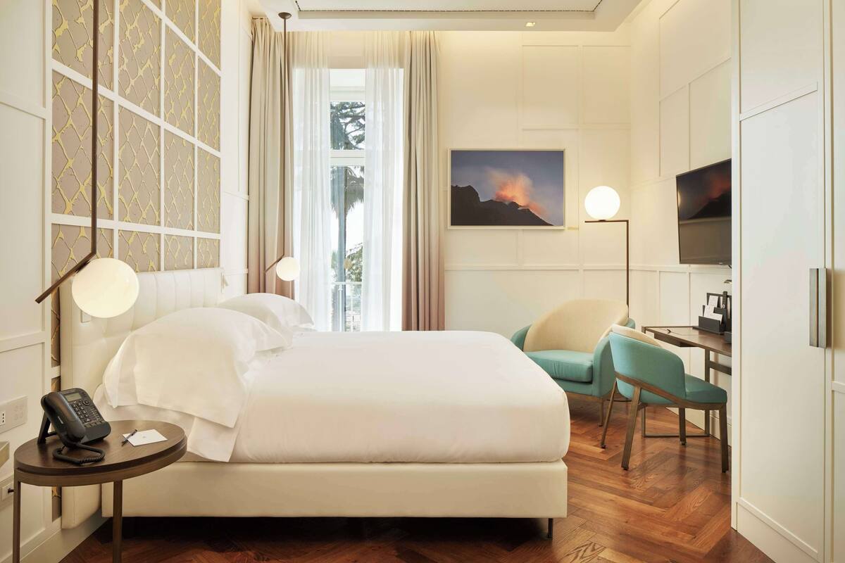 A hotel room with a bed, bedside table with a phone, a desk with a chair, a painting on the wall, and a window with curtains.