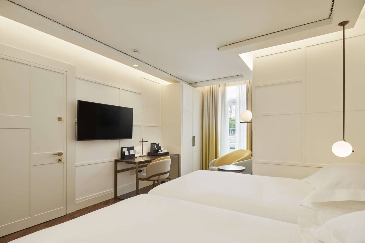A hotel room with a large bed, a desk with a chair, a flat-screen TV mounted on the wall, and a window with curtains.