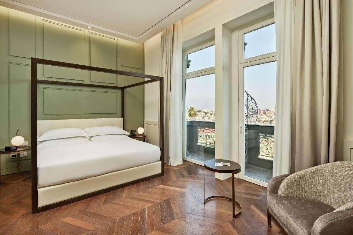A bedroom with a four-poster bed, a small table, a chair, and large windows with a view.