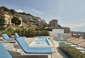 A rooftop pool area with lounge chairs, a hot tub, and a view of the cityscape.