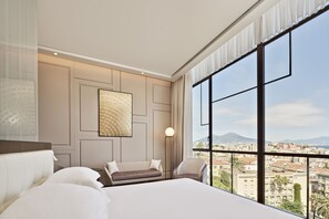 Een hotelkamer met een groot bed, een bank en een stoel. Er is een groot raam met uitzicht op een stadsbeeld en bergen.