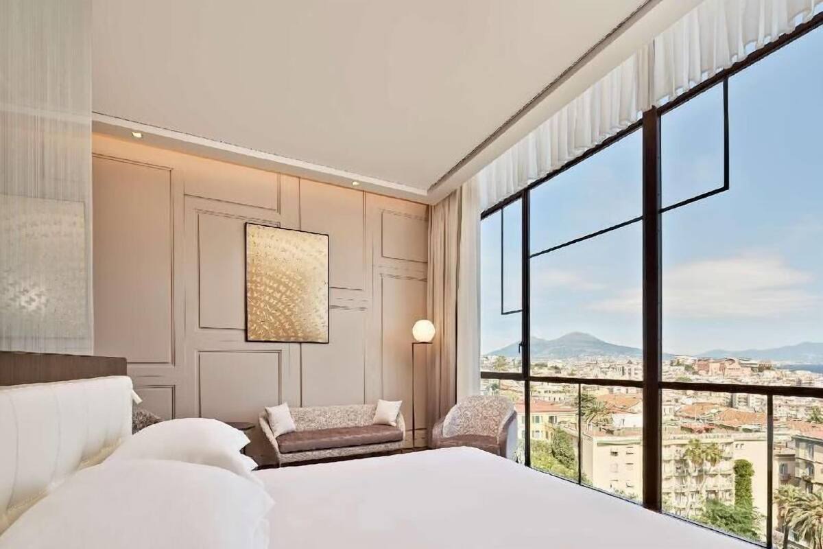 A hotel room with a large bed, a sofa, a chair, a wall-mounted artwork, and a city view through large windows.