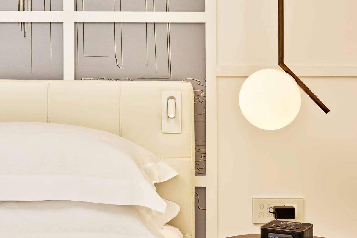 A hotel room with a bed, a bedside table with a digital clock, and a wall with framed architectural drawings.