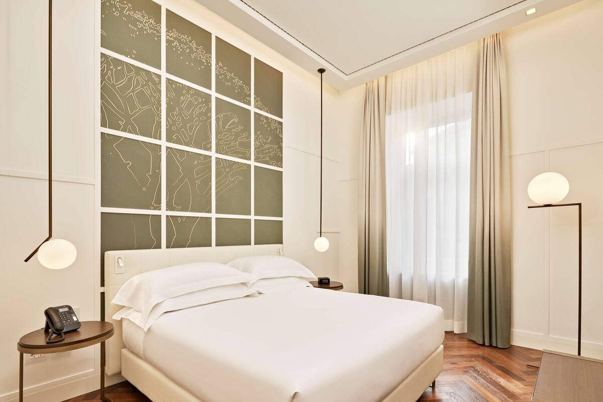 A modern hotel room with a large bed, a bedside table with a phone, a wall-mounted light, and a view of the city through a large window with a geometric pattern.