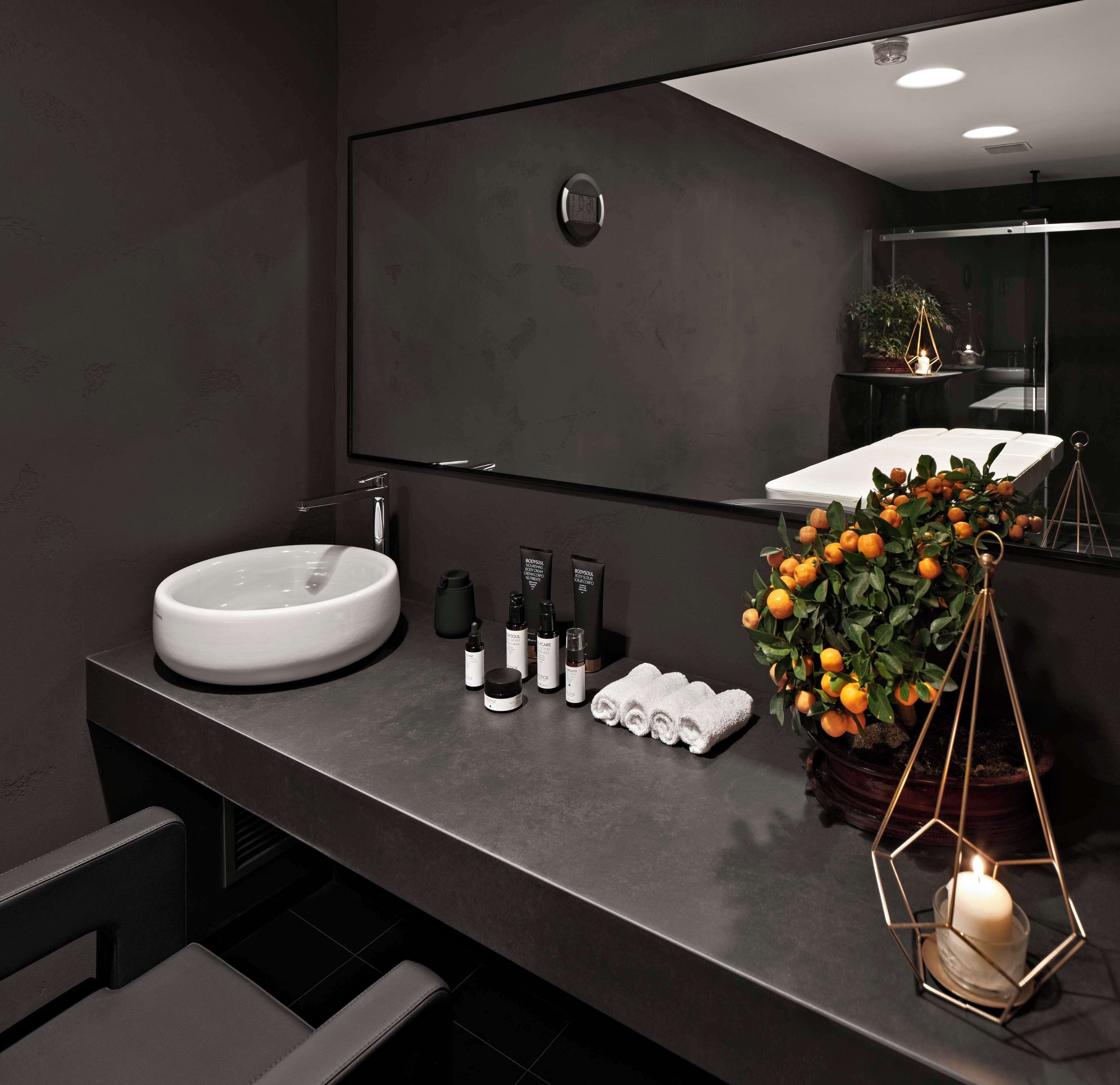 Een moderne badkamer met een grote spiegel, een rond wastafel en een donker aanrecht. Toiletartikelen, handdoeken en een potplant met mandarijnen zijn aanwezig.