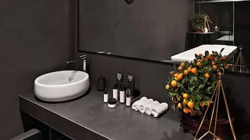 Een moderne badkamer met een grote spiegel, een rond wastafel en een donker aanrecht. Toiletartikelen, handdoeken en een potplant met mandarijnen zijn aanwezig.