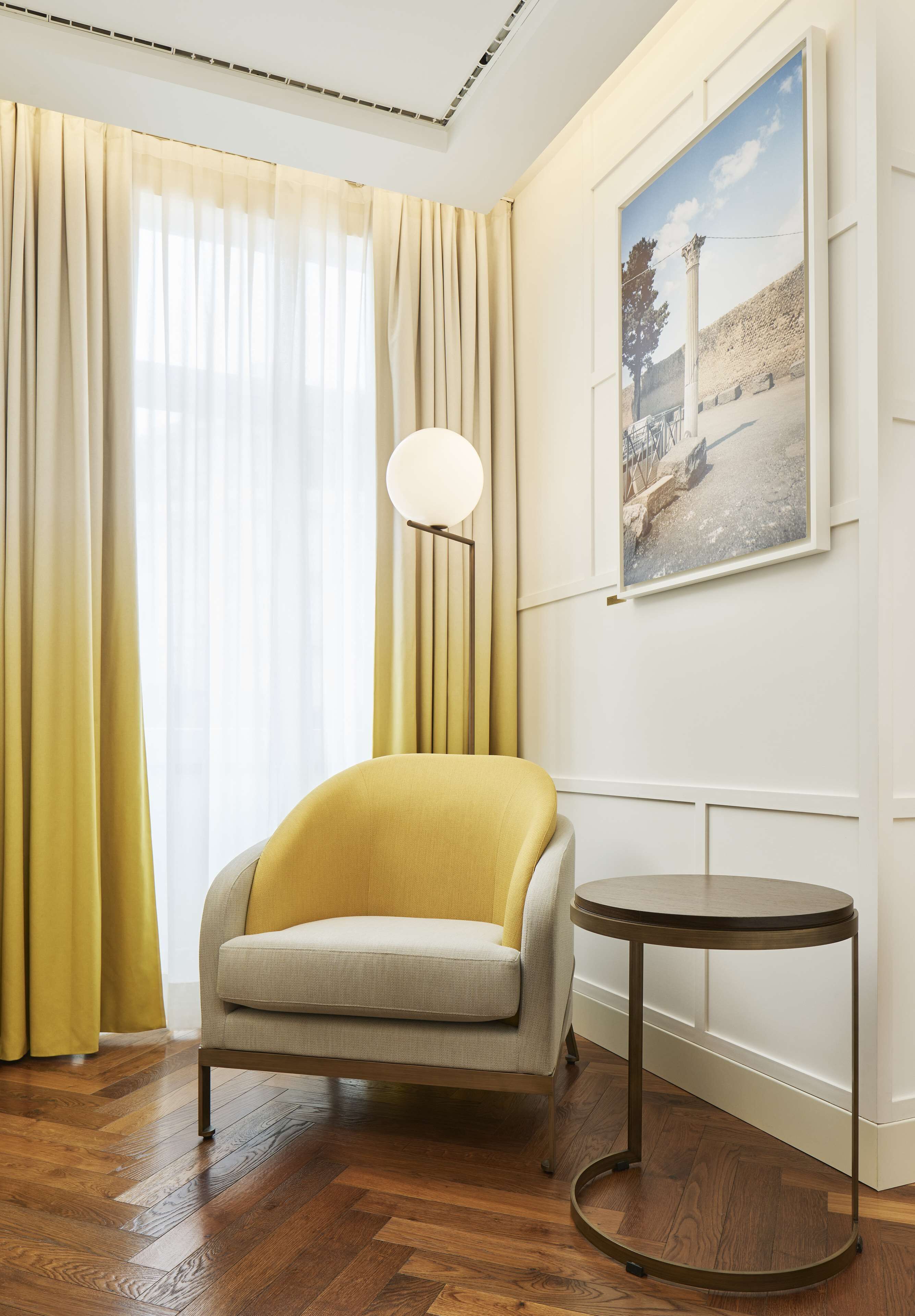 Una habitación con un sillón amarillo, una mesa redonda, un ventanal con cortinas translúcidas y un cuadro en la pared.
