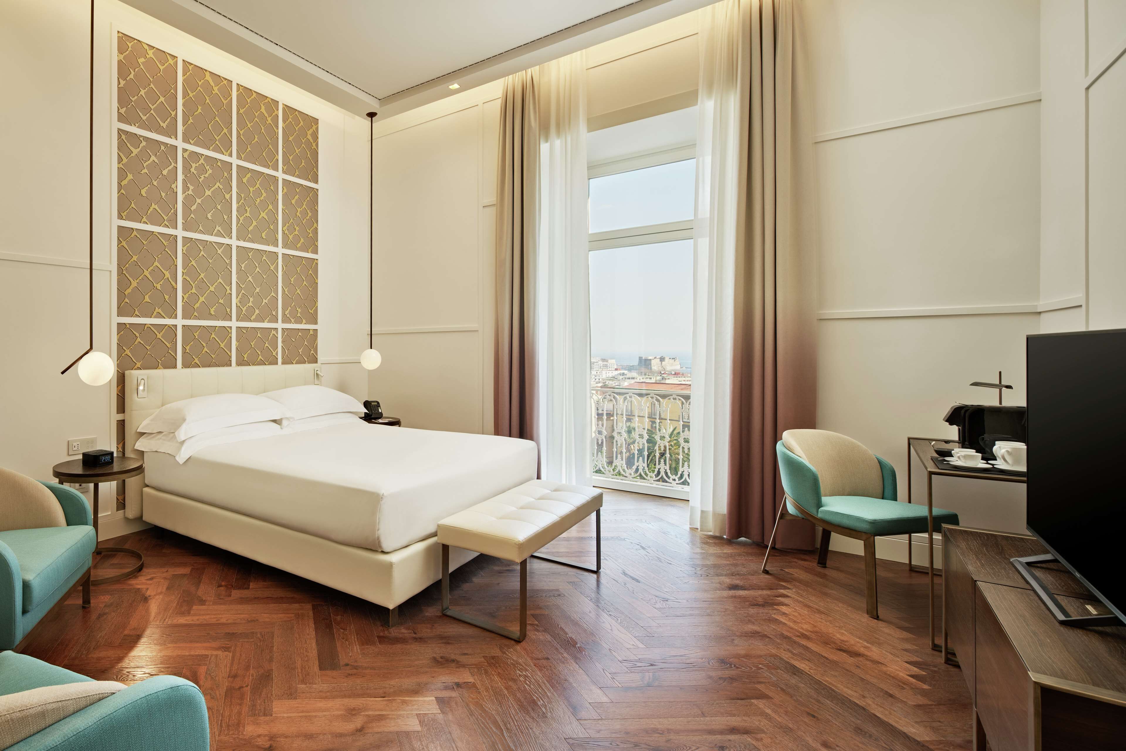 A hotel room with a large bed, a desk with a TV, a chair, and a view of the city through a window.