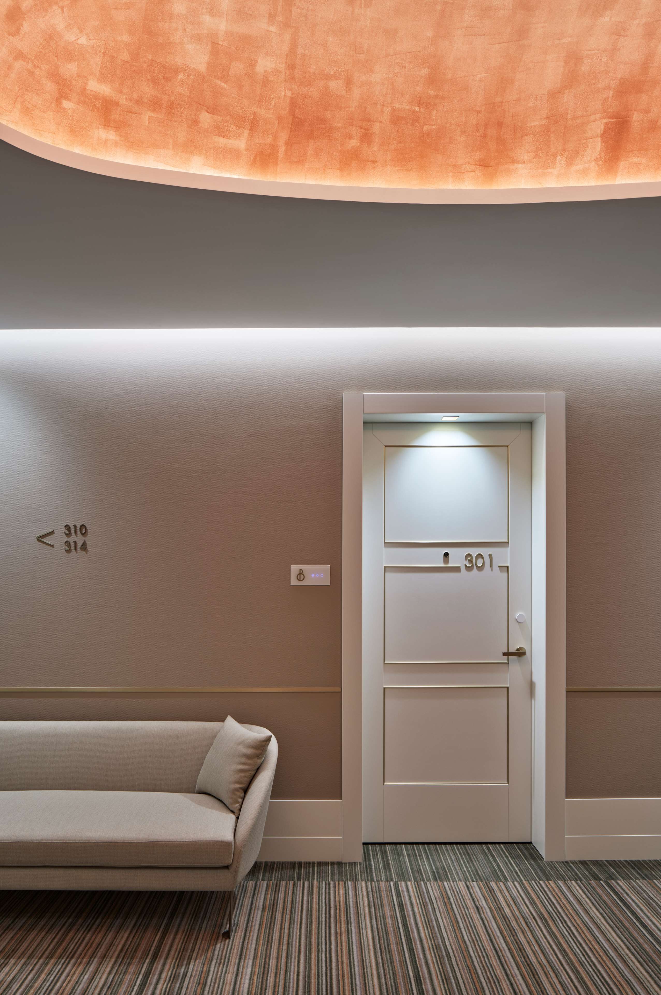 A hotel room with a sofa, a door with room numbers, and a carpeted floor.