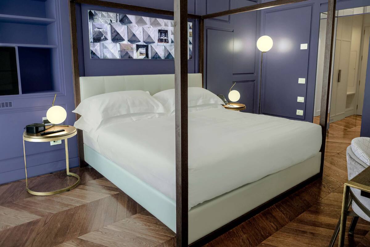 A neatly made bed with white linens, a bedside table with a lamp, and a wall with geometric artwork.