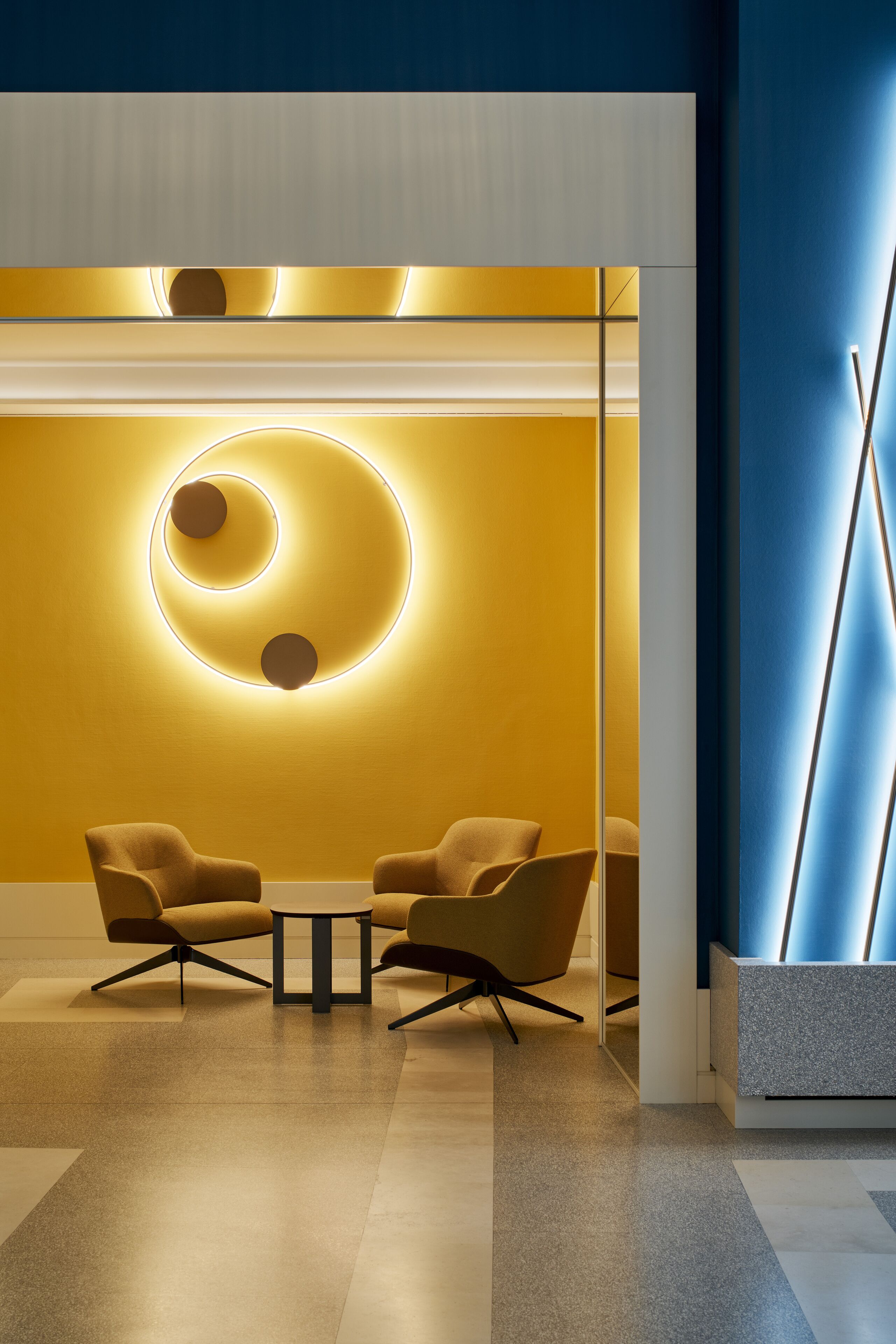 Un intérieur moderne avec un mur jaune, un luminaire circulaire et deux fauteuils disposés autour d’une petite table.