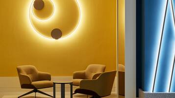 Un intérieur moderne avec un mur jaune, un luminaire circulaire et deux fauteuils disposés autour d’une petite table.