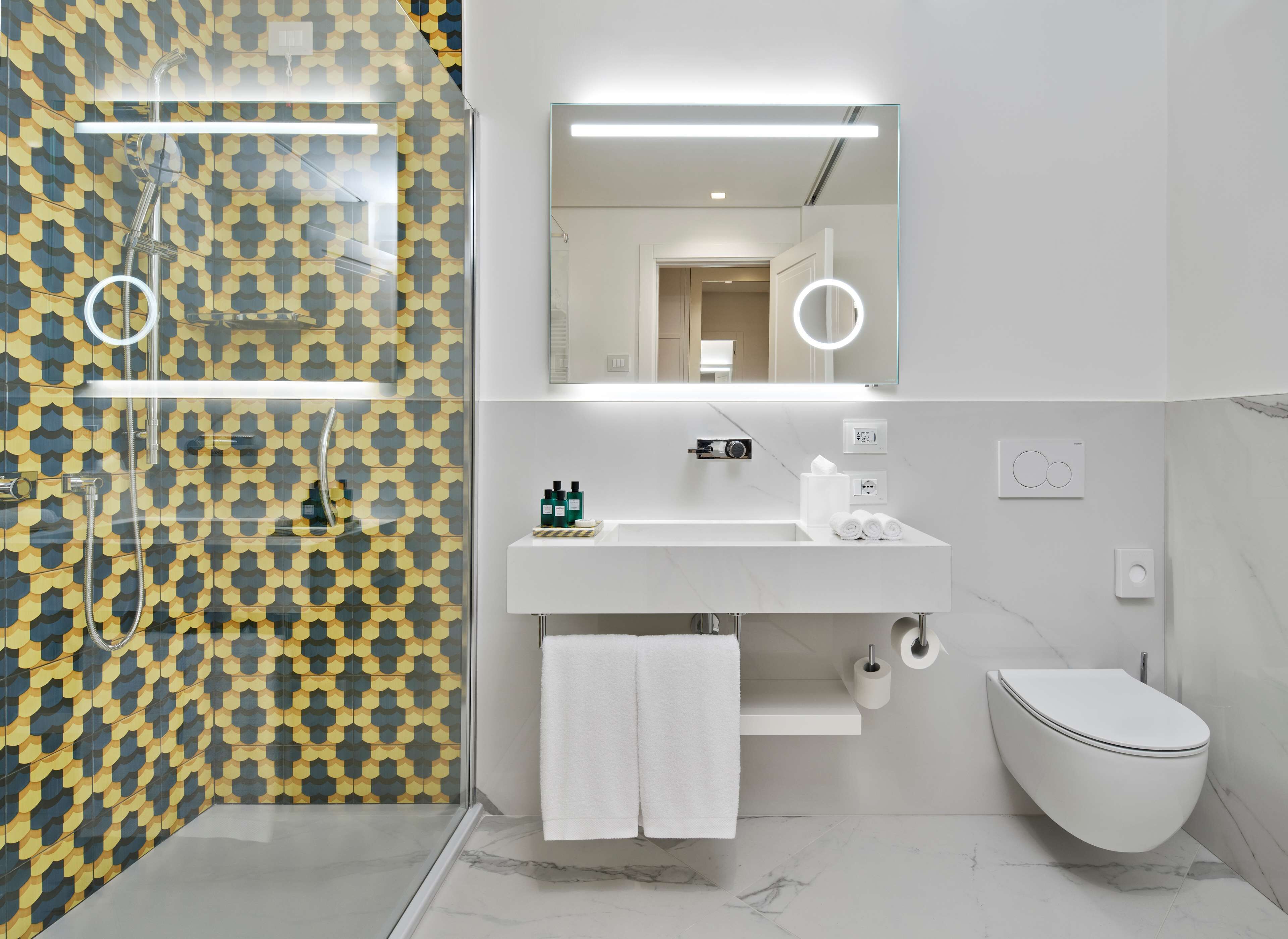 Un baño moderno con pared estampada, un lavabo blanco, un espejo con iluminación, un inodoro y una zona de ducha.