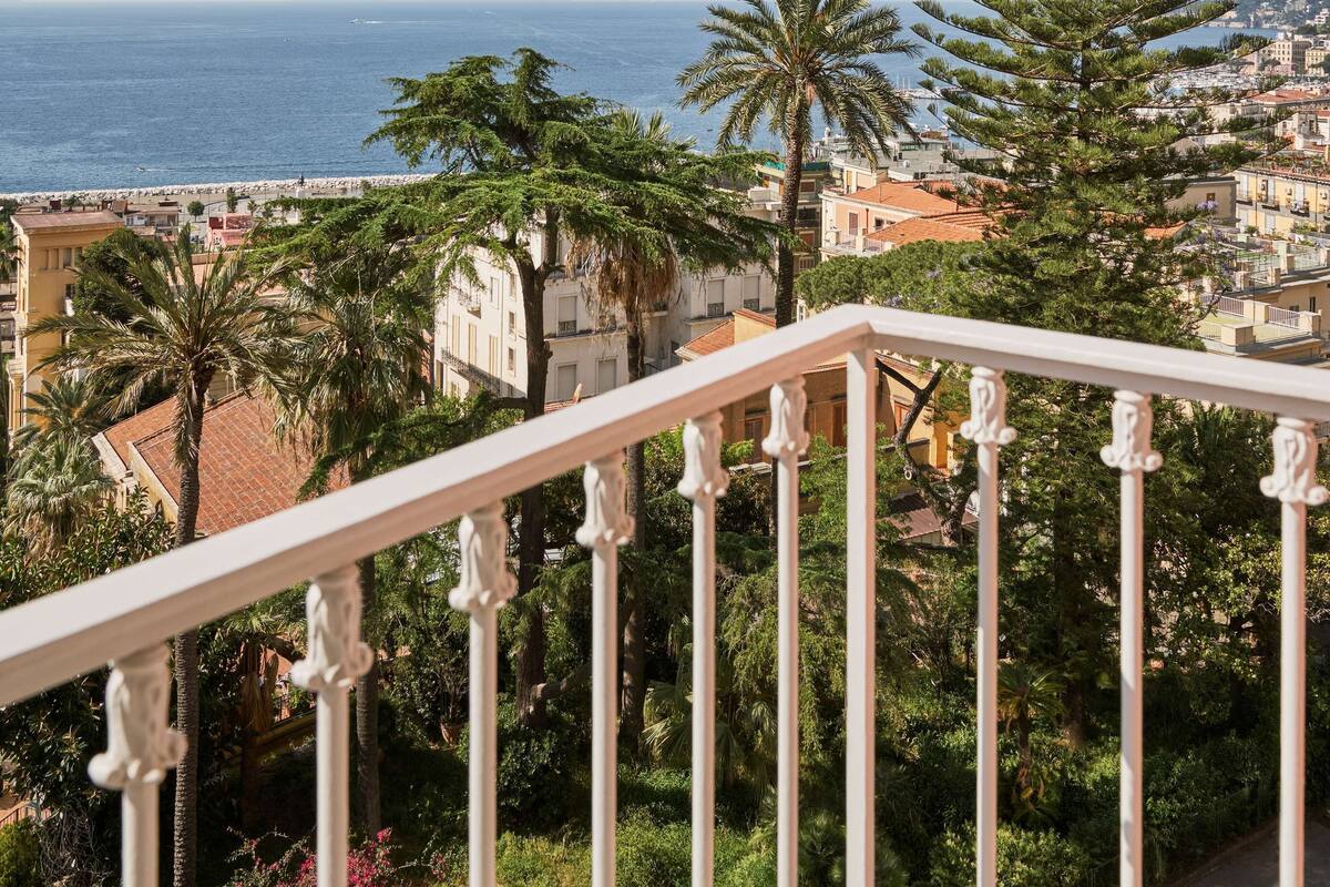 A balcony with a railing overlooking a coastal town, palm trees, and a view of the sea.