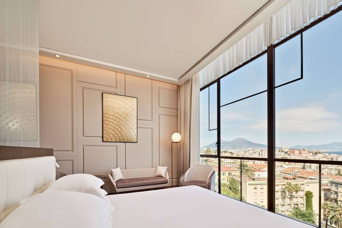 A hotel room with a large bed, a sofa, a chair, and a large window offering a city view.