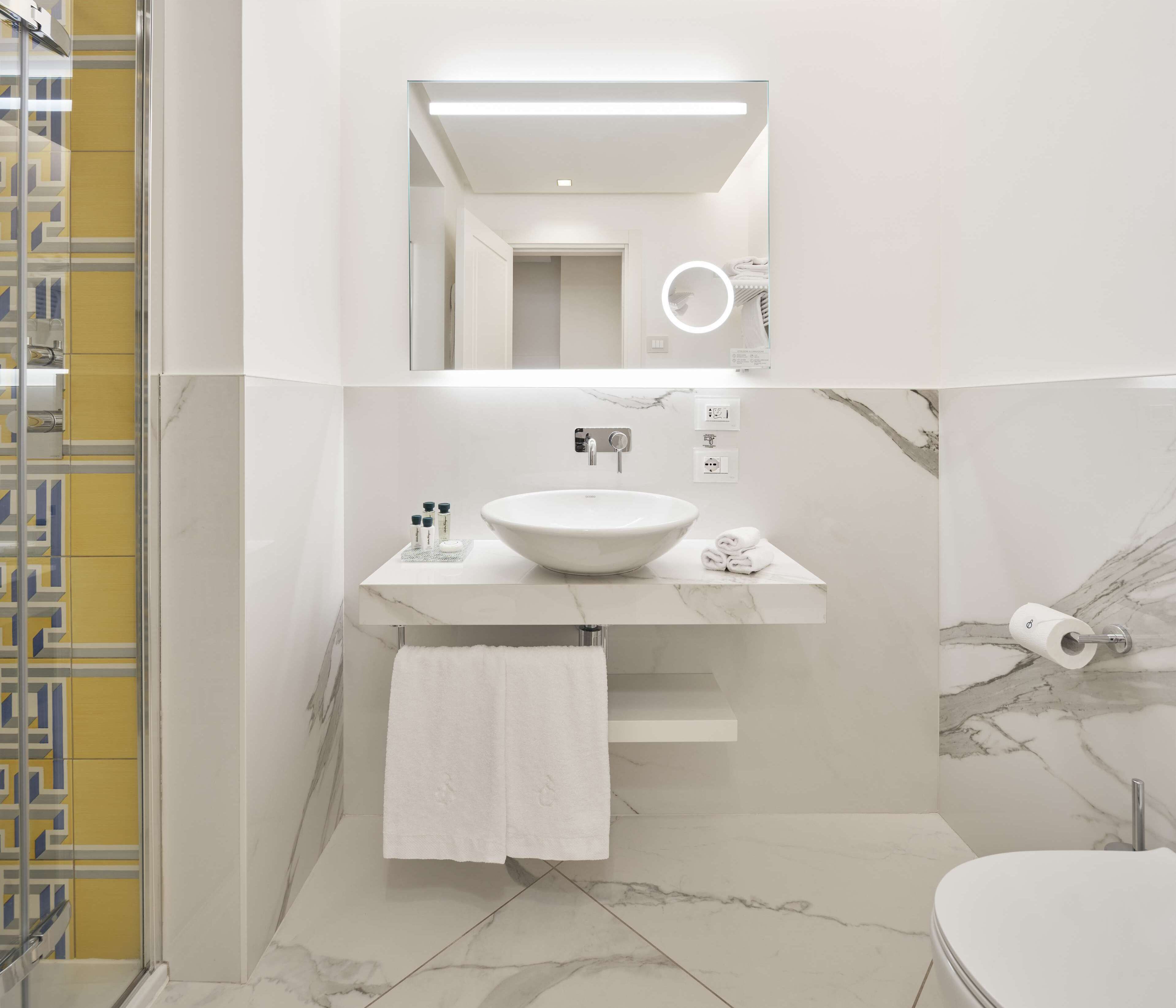 A modern bathroom with a large marble sink, a mirror with a circular light, and a towel hanging on a rack.
