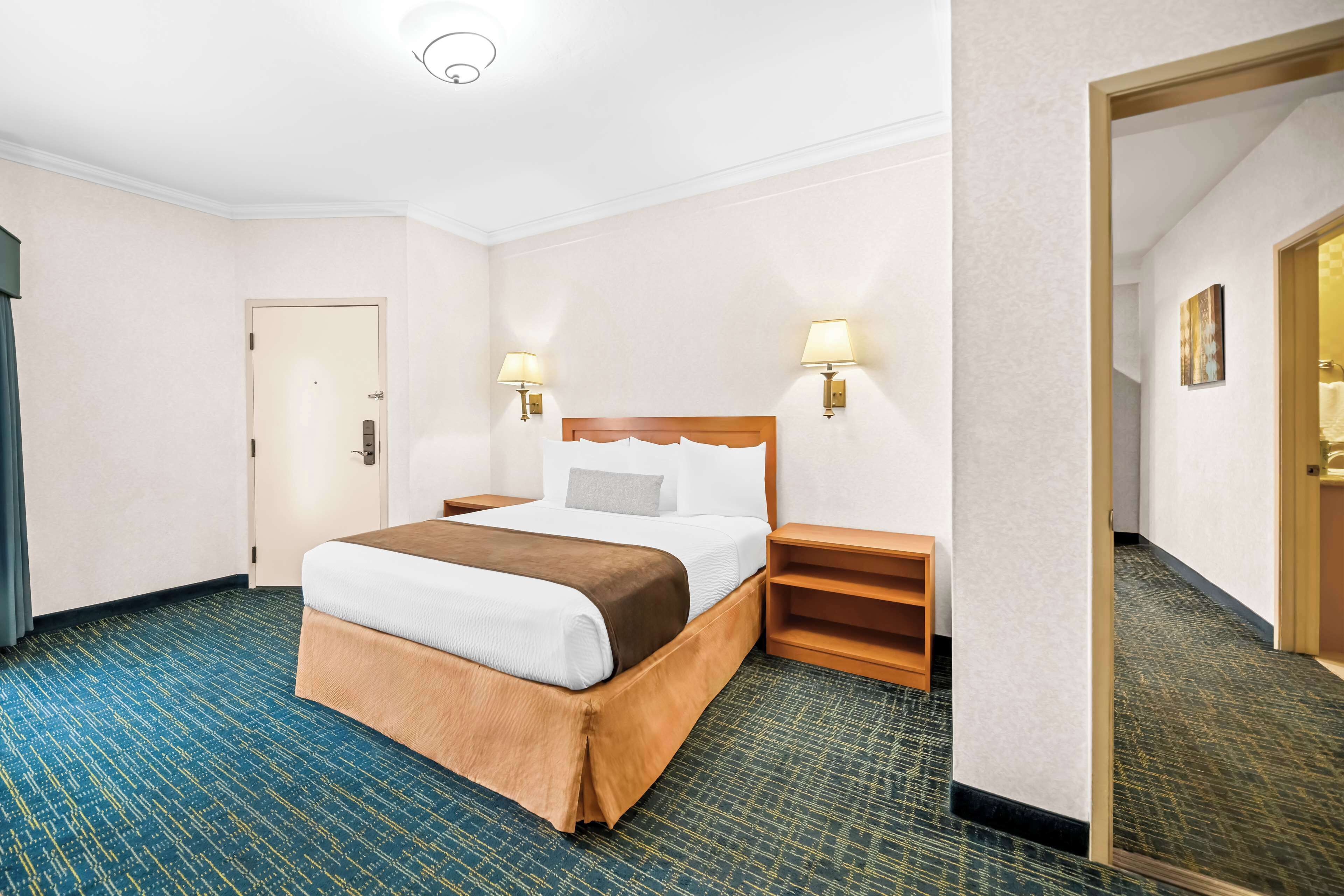 A hotel room with a large bed, two bedside tables with lamps, a wooden headboard, and a carpeted floor.