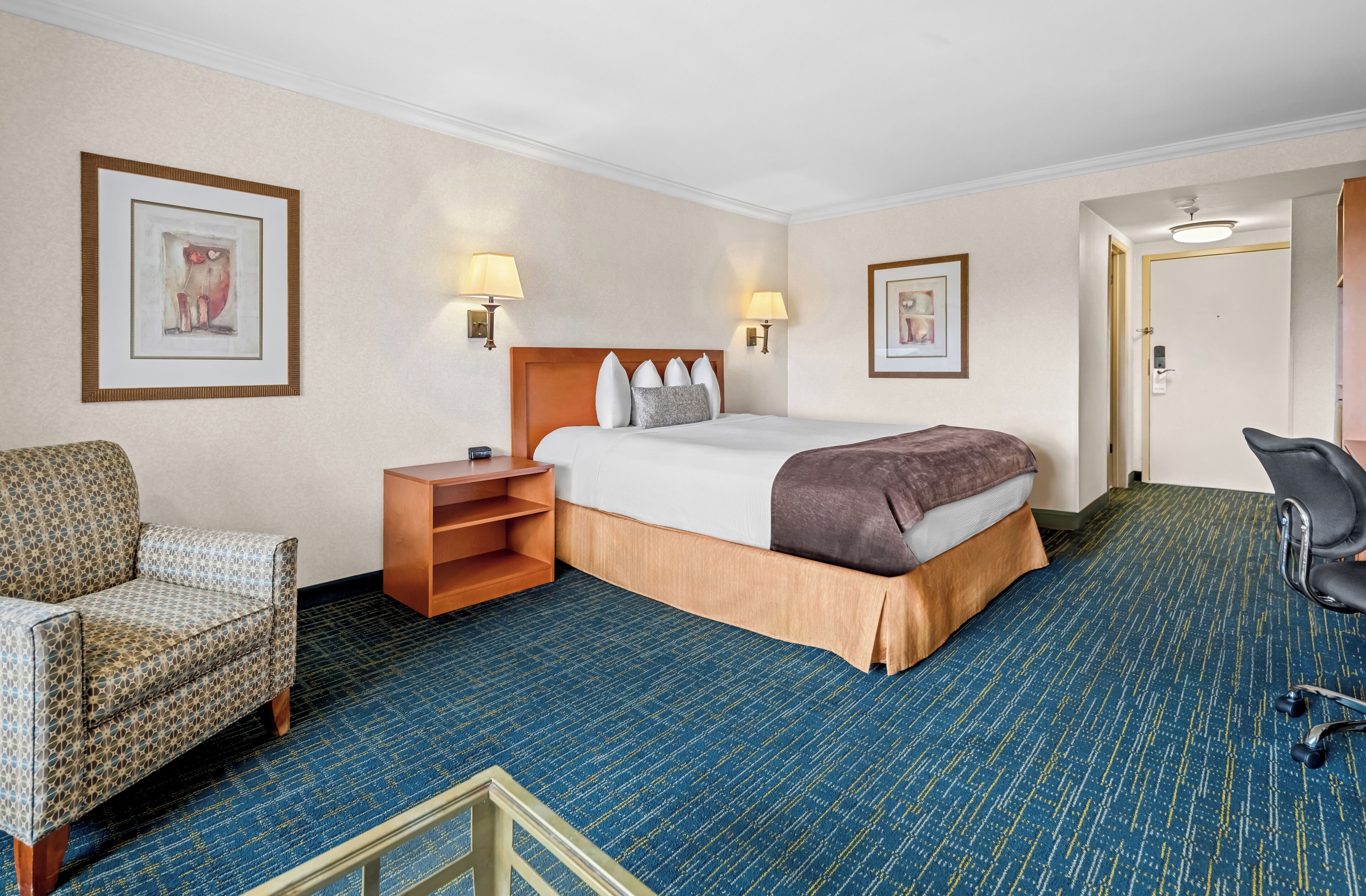A hotel room with a bed, bedside table, lamp, chair, and carpet.
