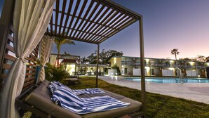 Eine Poollounge mit überdachter Sitzgruppe, einem Pool und einem Gebäude mit mehreren Balkonen im Hintergrund.