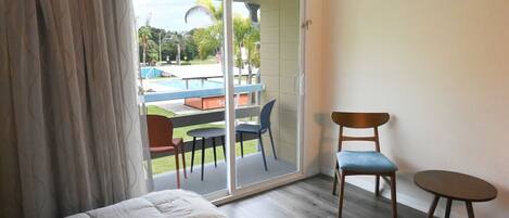 Habitación de hotel con una cama, una mesita, una silla y vistas a una piscina y zona de estar exterior a través de un ventanal corredero.