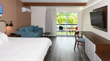 A hotel room with a bed, a blue sofa, a desk with a chair, a TV, and a balcony with a view of greenery.