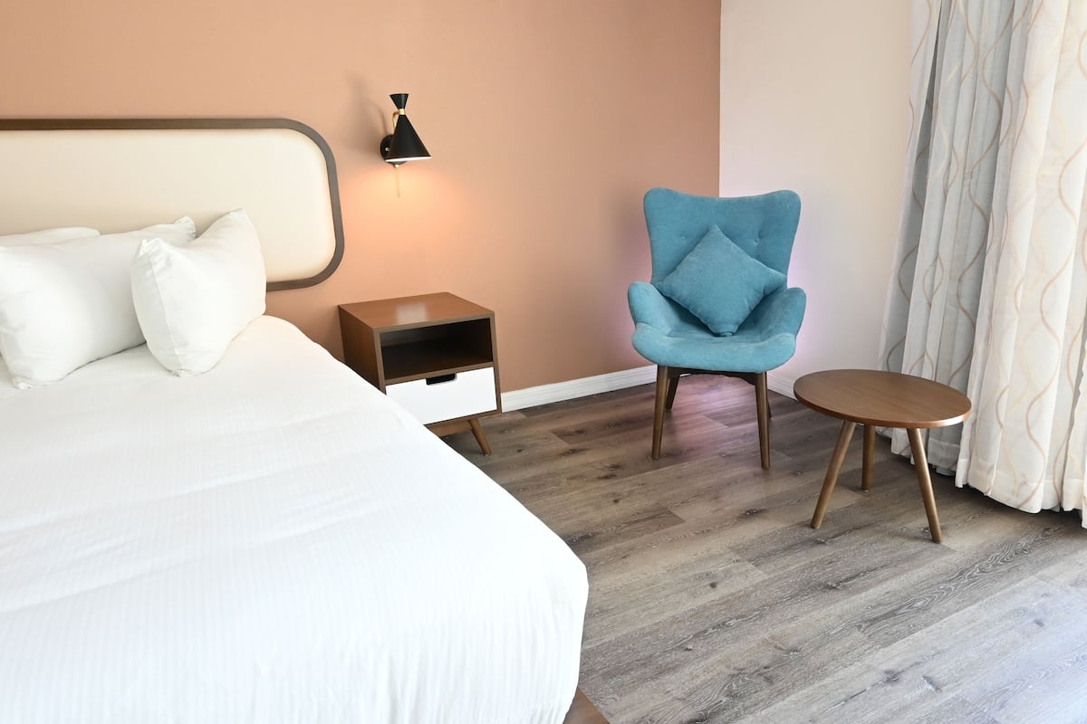 A hotel room with a bed, a nightstand, a lamp, a blue armchair, and a small table.