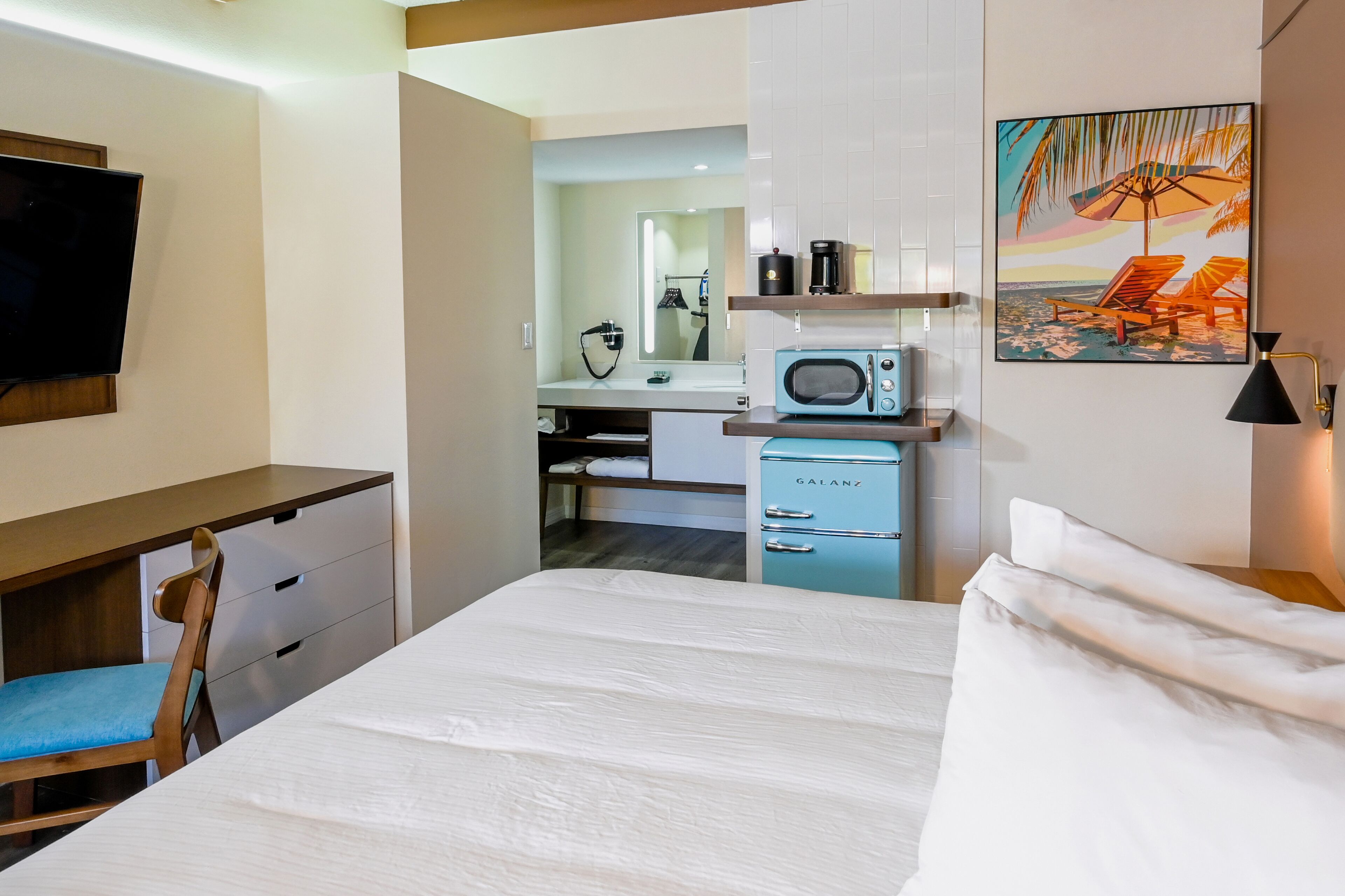 Habitación de hotel con cama, escritorio y silla, televisor, microondas, cafetera, minibar y un cuadro de una playa.