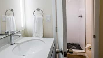 A bathroom with a white sink, two towel rings, a shower with a black patterned floor, and a toilet with a paper towel dispenser.