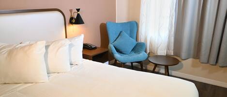 Une chambre d’hôtel avec un grand lit, un fauteuil bleu, une petite table ronde, une lampe de chevet et une fenêtre avec des rideaux.