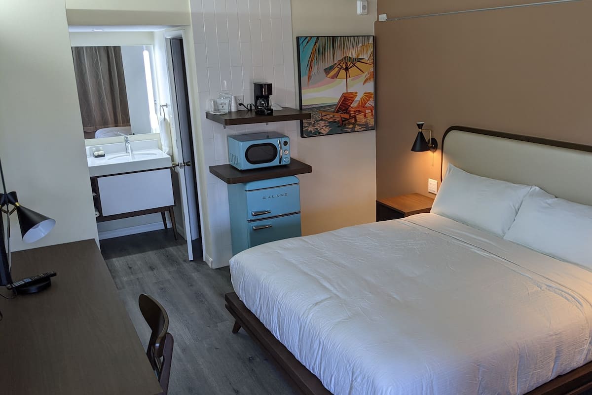 A hotel room with a bed, a desk with a chair, a microwave, a coffee maker, and a painting on the wall.