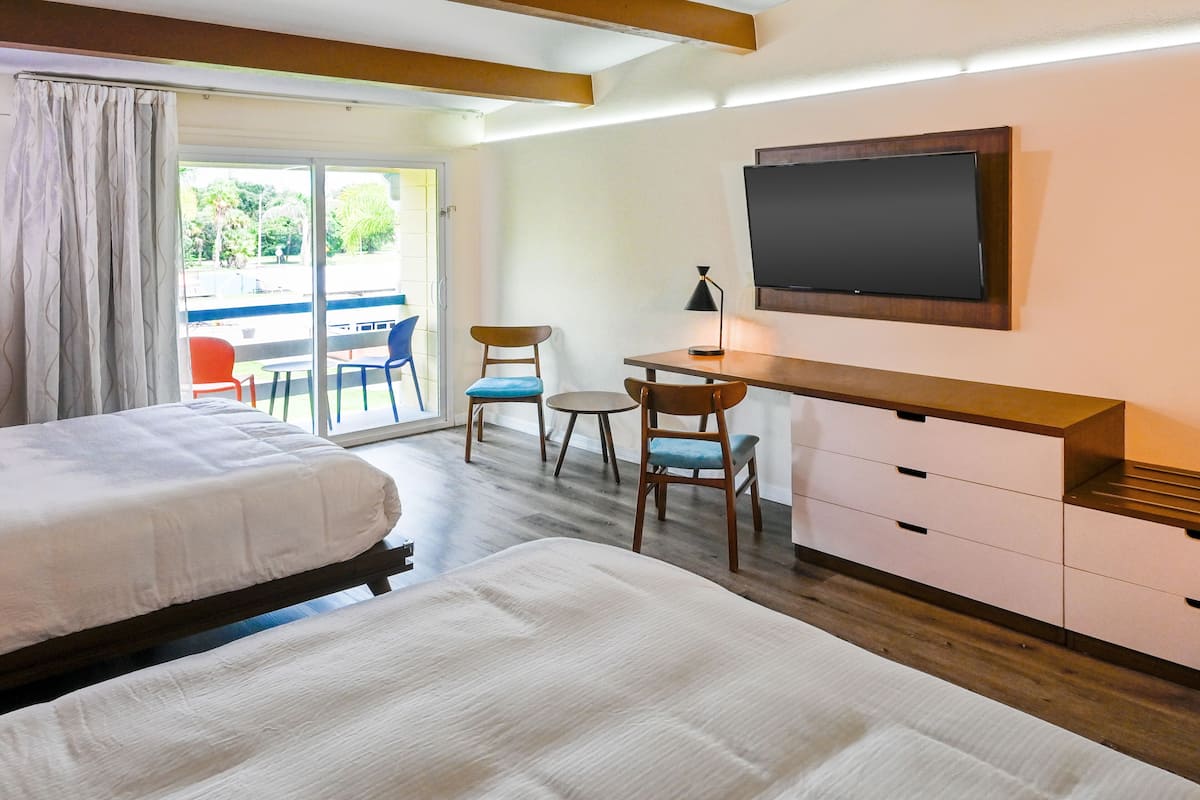 A hotel room with two beds, a desk, a chair, a TV, and a sliding glass door to the outside.