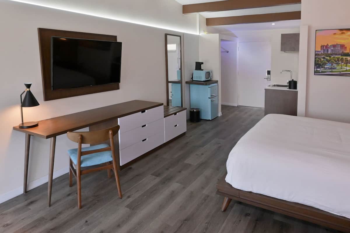 A bedroom with a bed, a desk with a chair, a TV, a mirror, a kitchenette, and a bathroom.