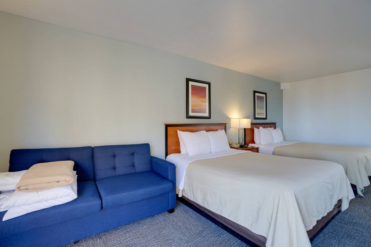Une chambre d’hôtel avec un canapé bleu, deux lits simples, une tête de lit en bois et un tableau encadré au mur.