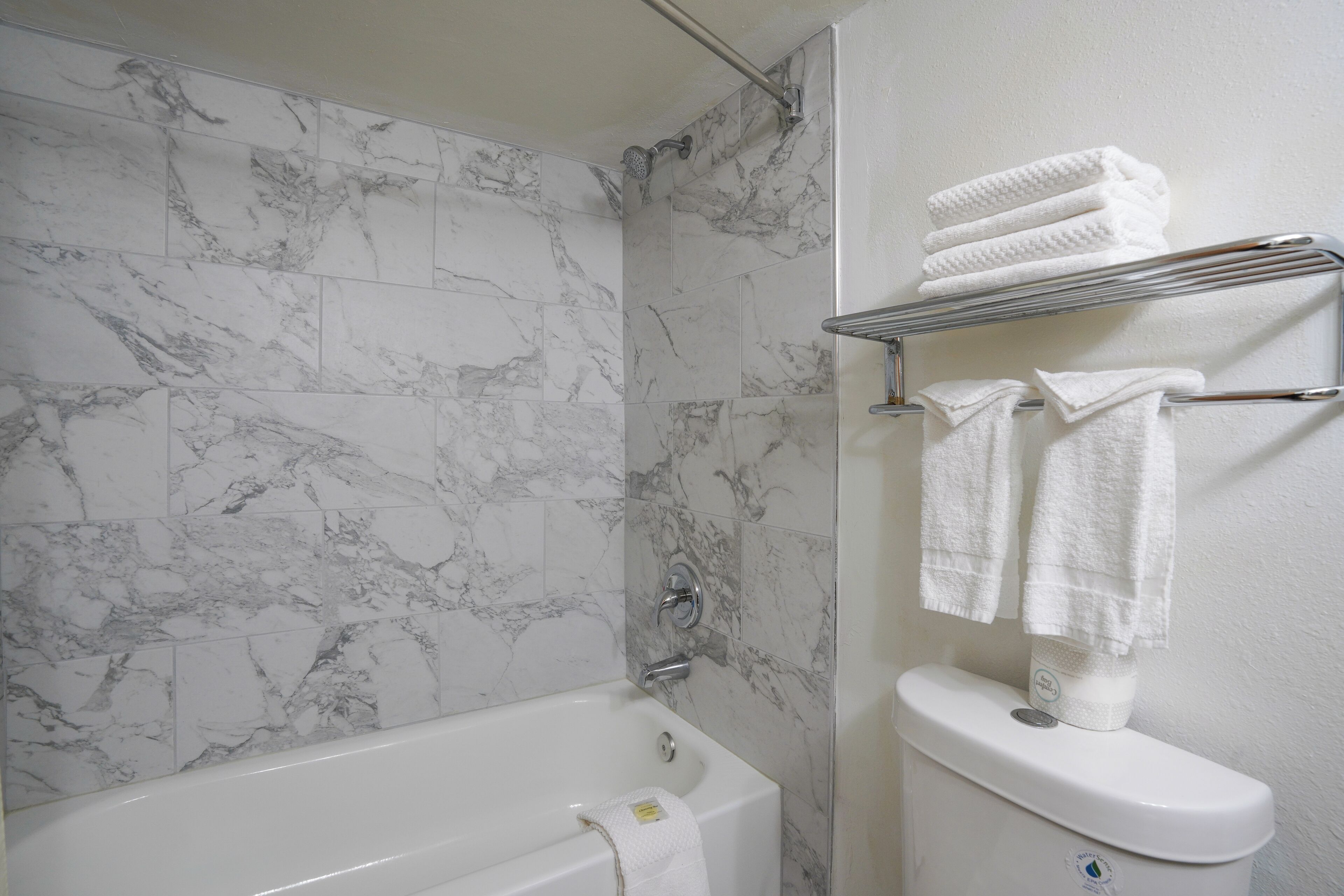 Un baño con pared de ducha de azulejos de mármol, una bañera blanca y un inodoro con un perchero encima.