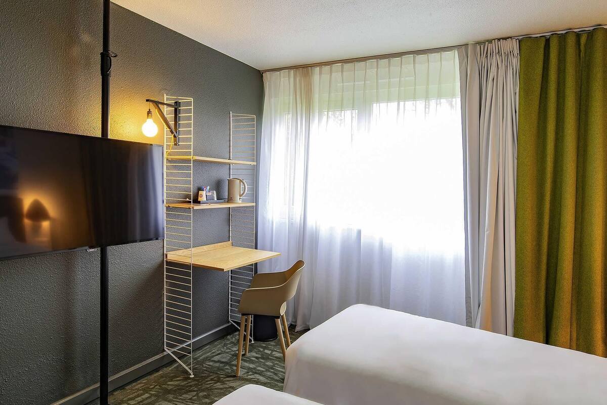 Une chambre d’hôtel avec un lit, un bureau, une chaise, une télévision et une fenêtre avec des rideaux.