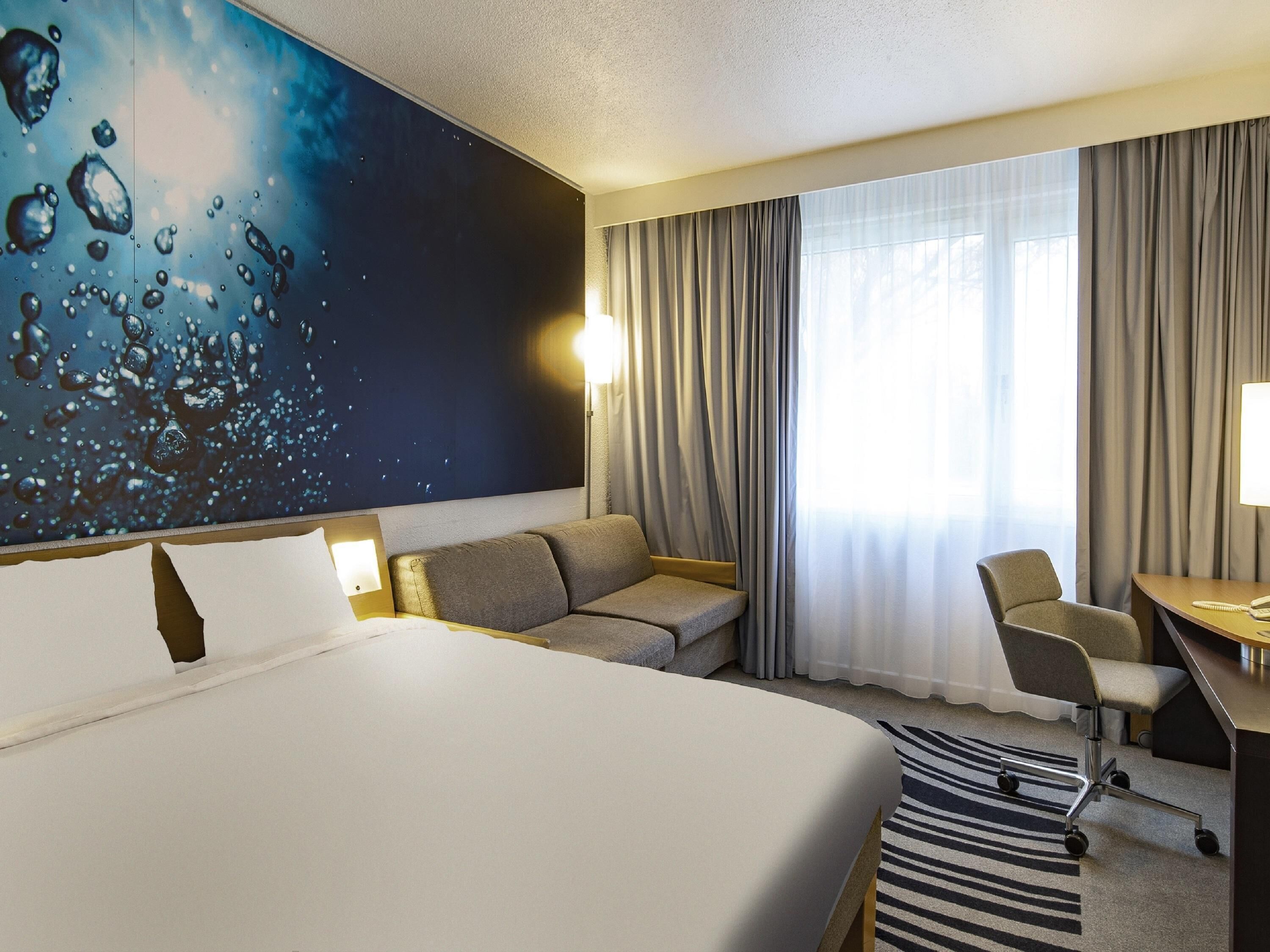 Una camera d'albergo con un letto grande, un divano, una scrivania, una sedia e un'ampia finestra con tende.
