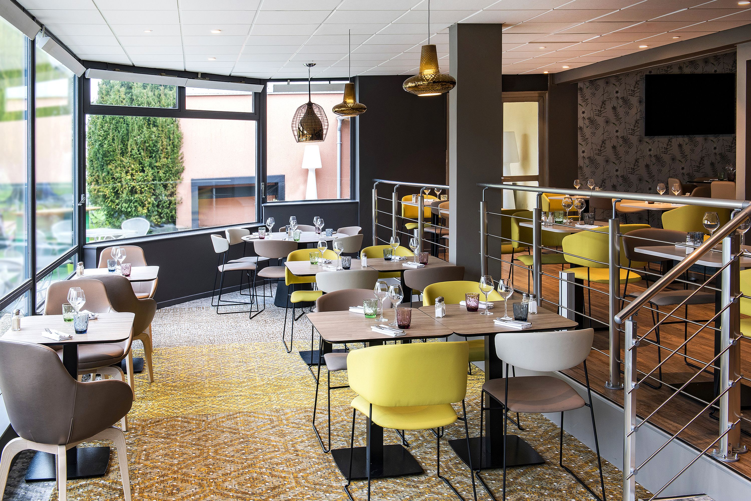 A modern restaurant with yellow and brown chairs, tables set with glasses and cutlery, and a large window offering a view of greenery.