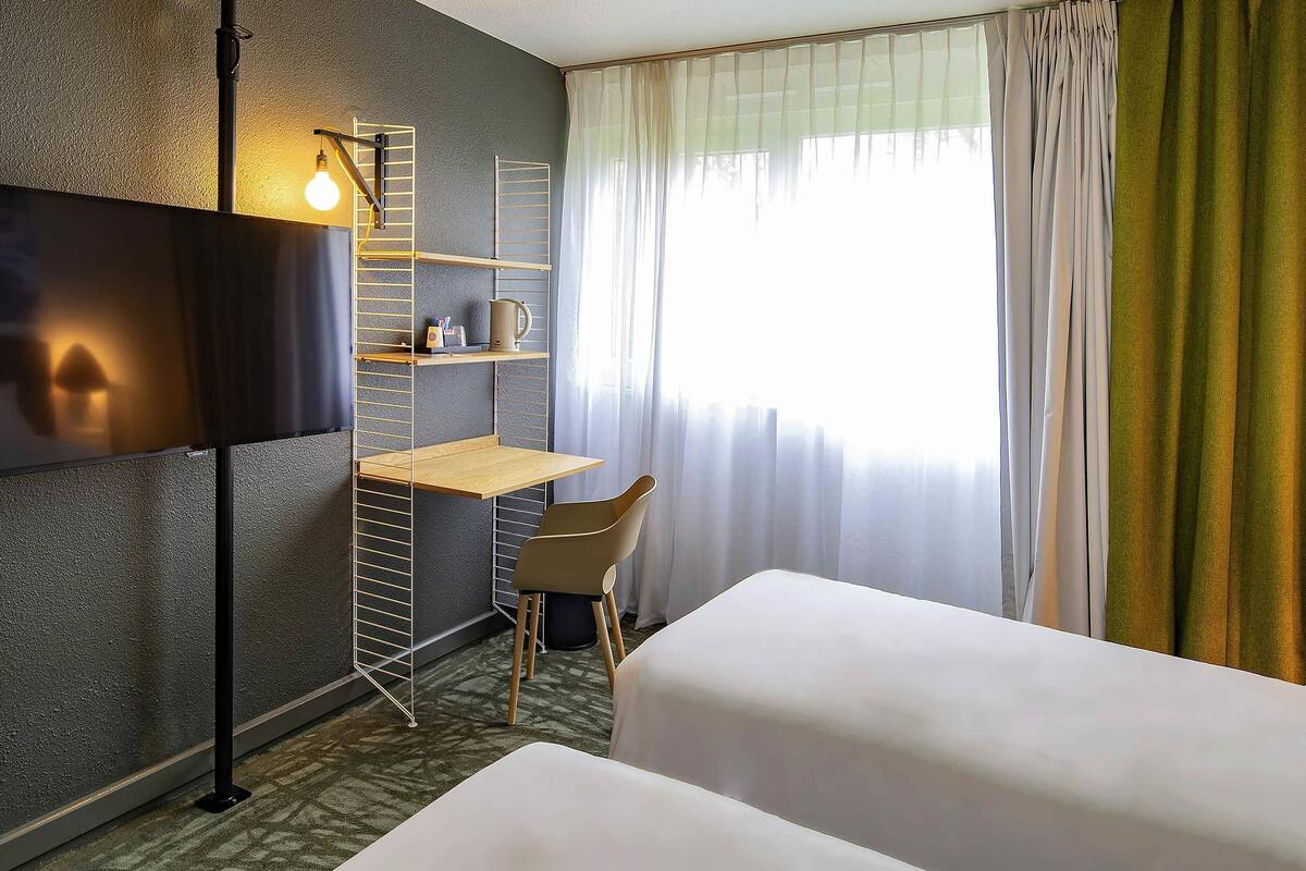A hotel room with a bed, a desk, a chair, a TV, and a window with curtains.