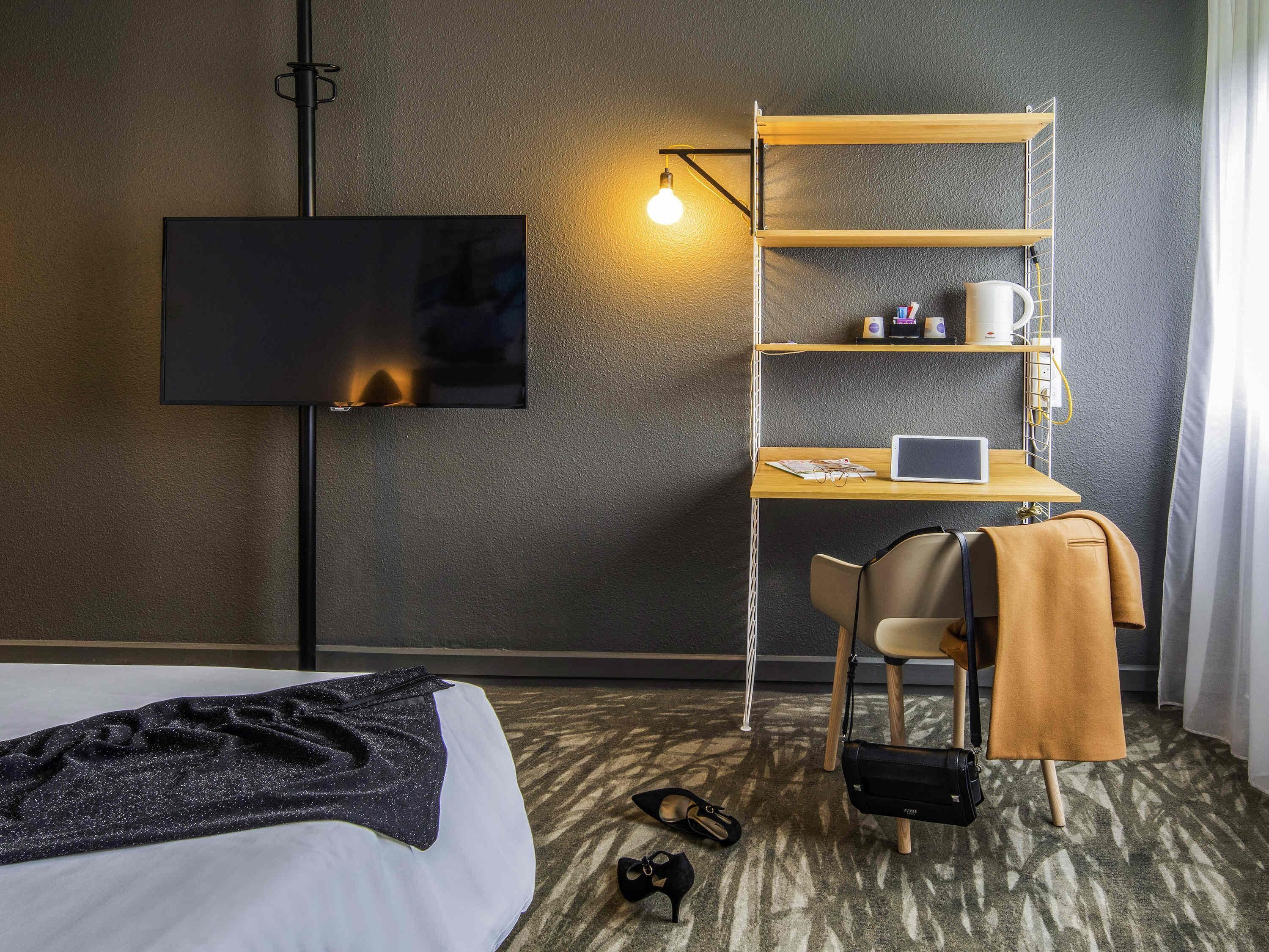 A hotel room with a bed, a desk with a laptop, a TV, and a shelf with items.