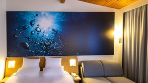 A hotel room with a large bed, a grey armchair, a nightstand with a phone, and a wall-mounted artwork depicting water droplets. - Novotel Metz Amneville Hotel (Hauconcourt)