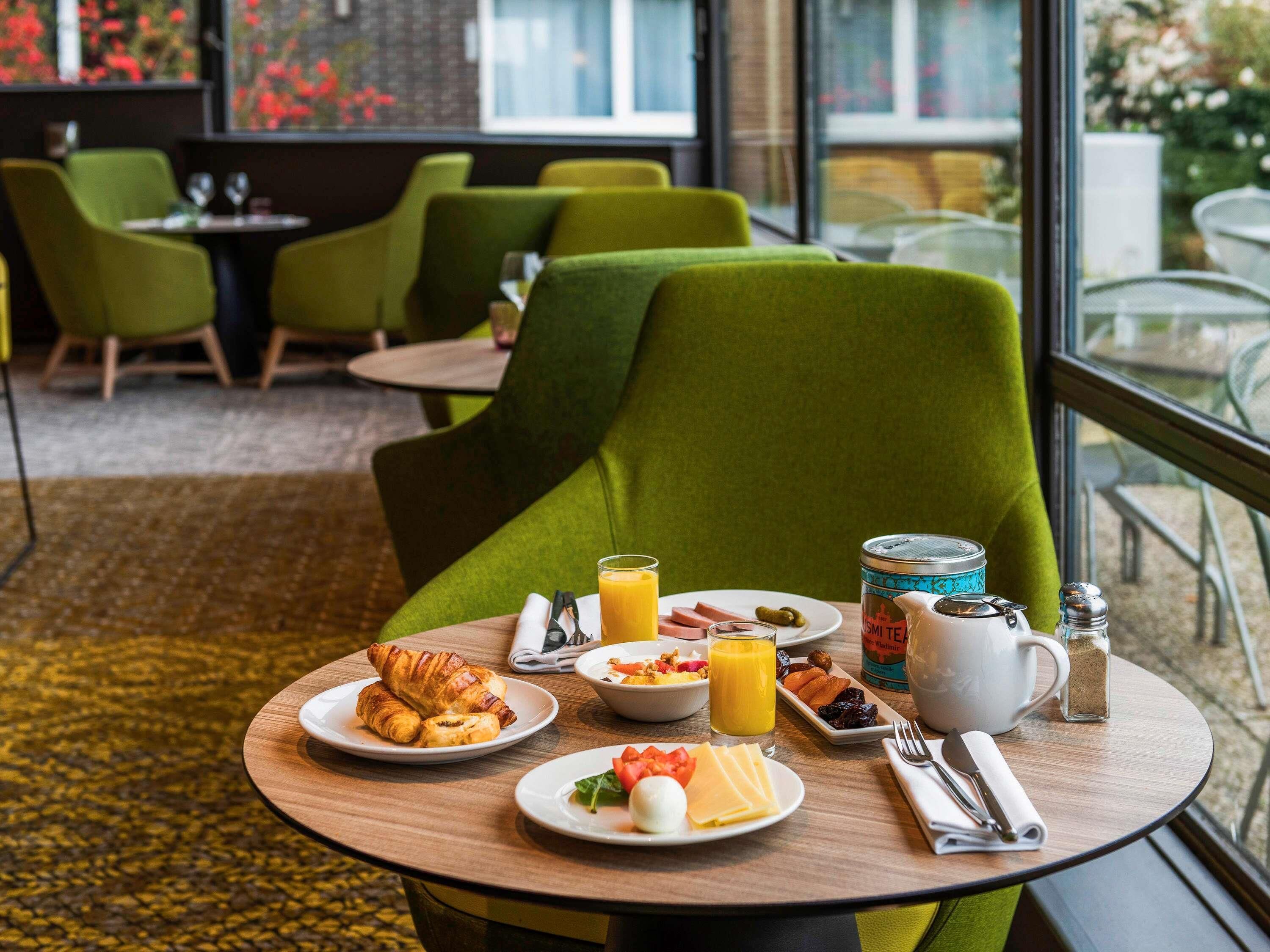 A breakfast setting with croissants, fruit, and juice on a wooden table, green chairs, and a view of the outdoors.