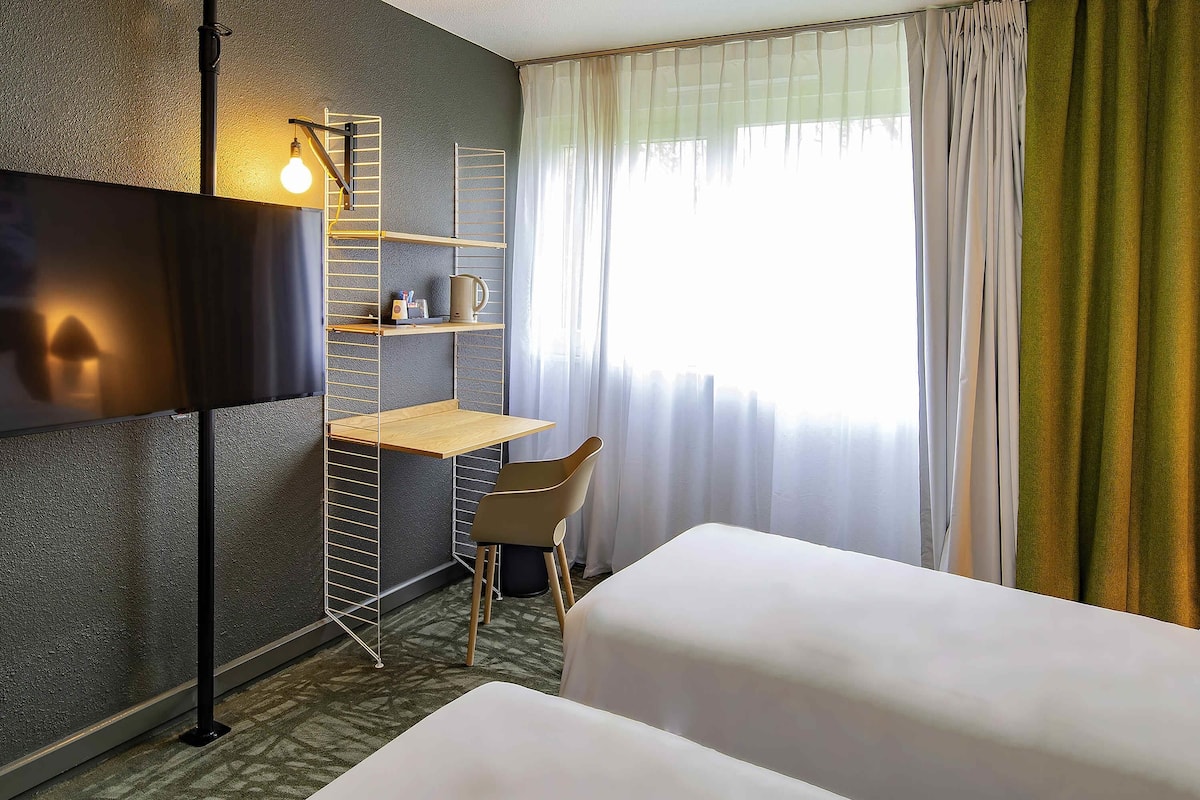 Une chambre d’hôtel avec un lit, un bureau, une chaise, une télévision et une fenêtre avec des rideaux.