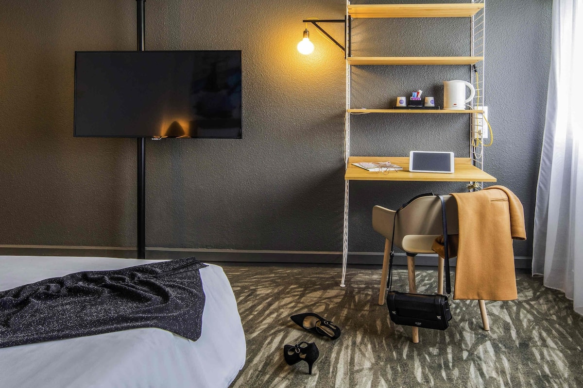 A hotel room with a bed, a desk with a laptop, a TV, and a shelf with items.
