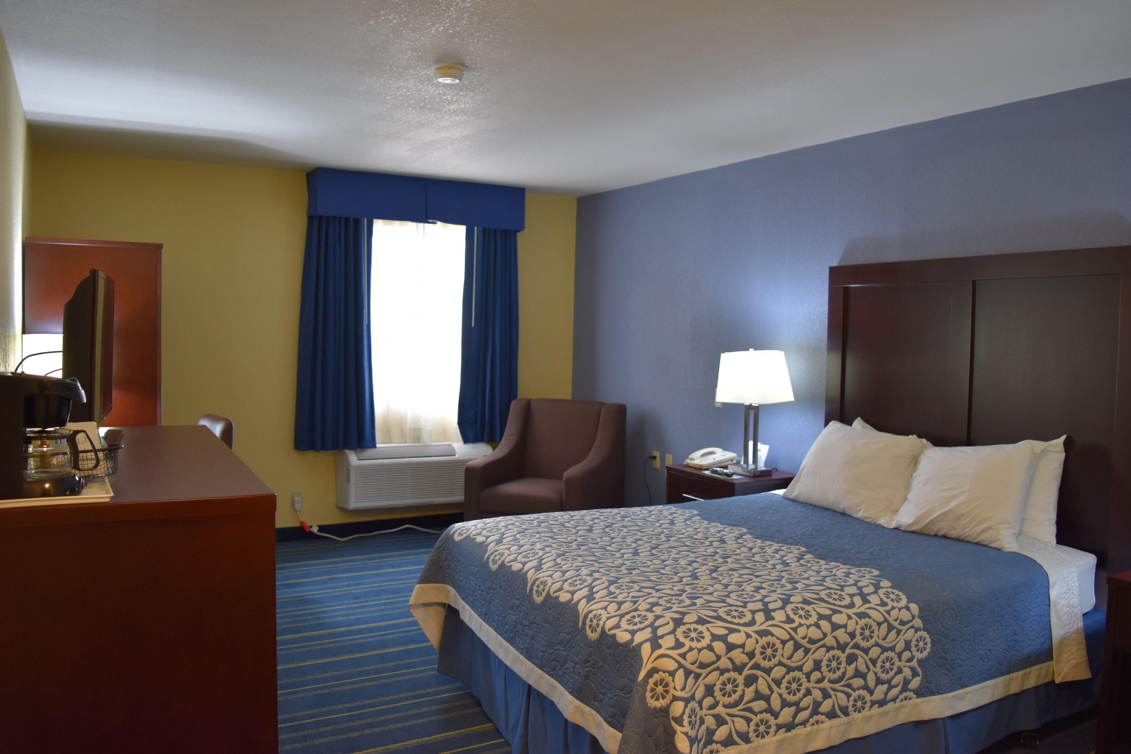 Una habitación de hotel con cama, escritorio, silla, lámpara y ventana con cortinas.