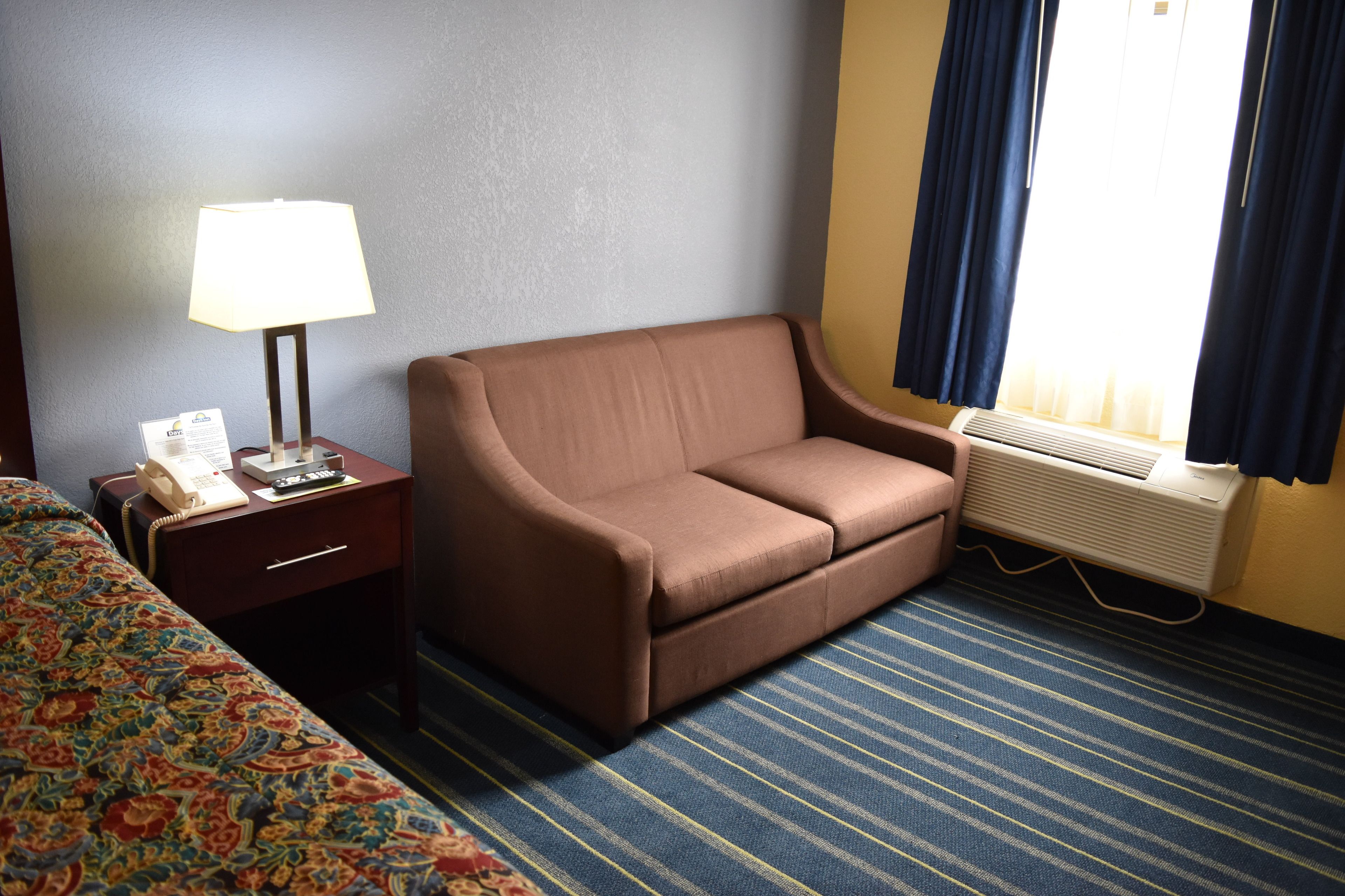 A hotel room with a brown sofa, a bedside table with a lamp, a telephone, and a window with curtains.