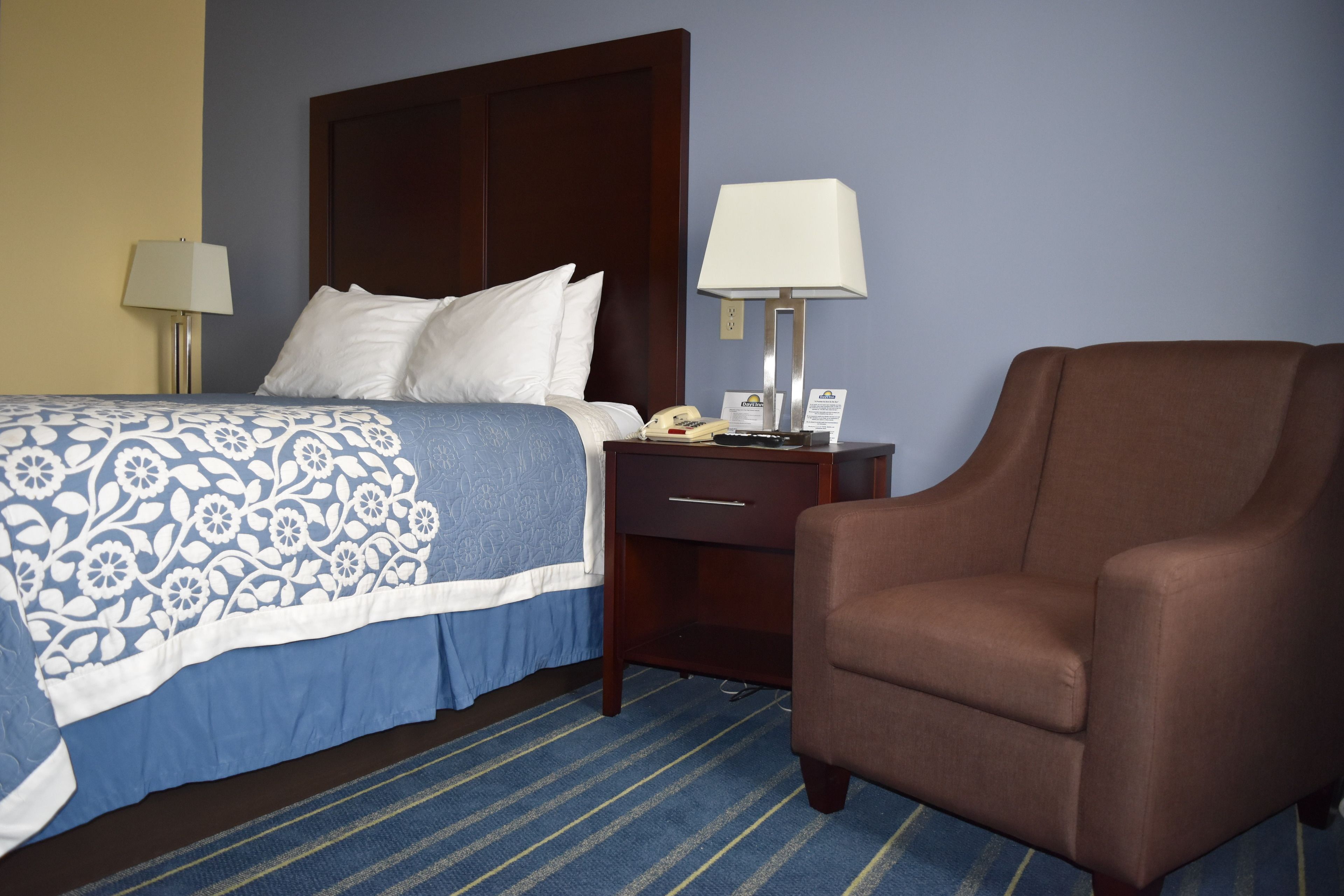 Une chambre d’hôtel avec un lit, deux lampes de chevet, un fauteuil marron, une table de chevet et une couvre-lit à motifs bleus et blancs.