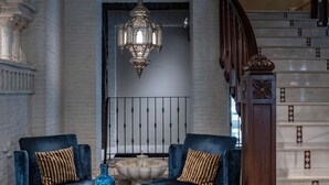 Amplia sala de estar con dos sofás azules, una mesa de centro redonda, una gran araña y una escalera con barandilla de madera.