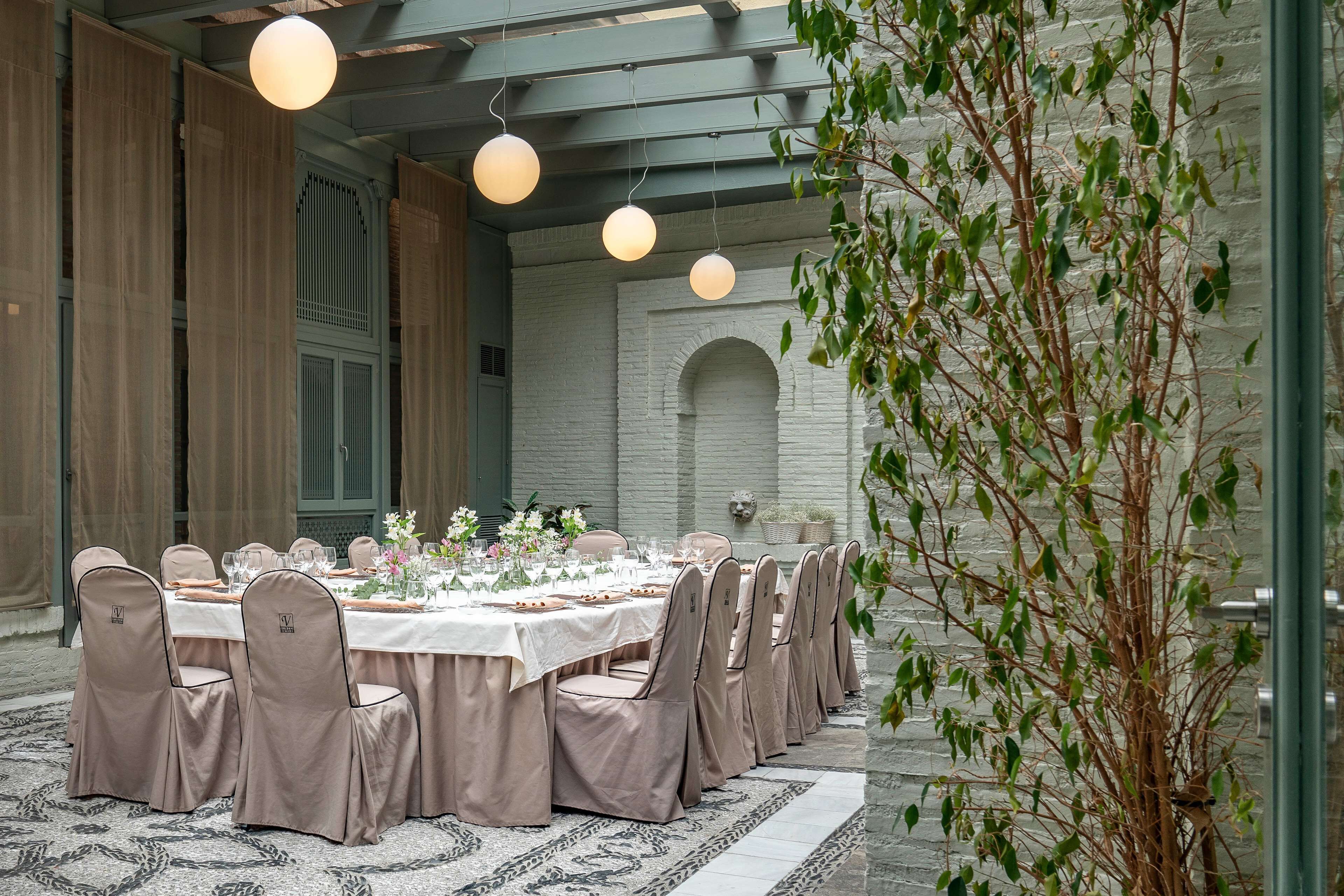 A formal dining room with a long table set for a meal, surrounded by chairs with headrests, and a large potted plant to the right.