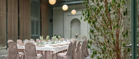 A formal dining room with a long table set for a meal, surrounded by chairs with headrests, and a large potted plant to the right.